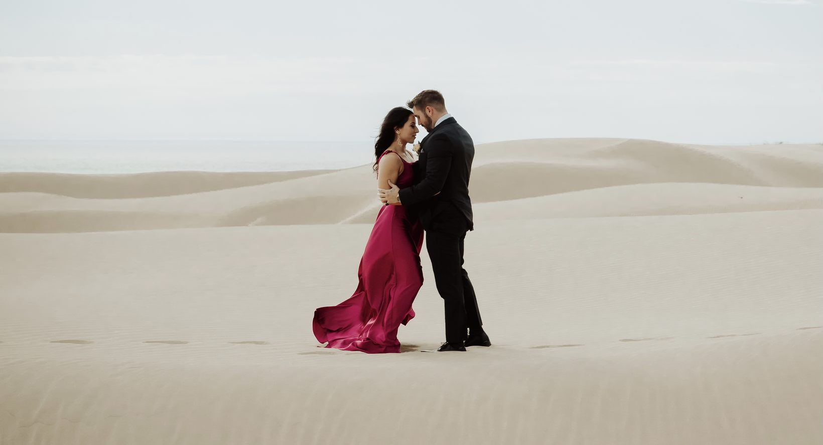 A couple embracing on open sand dunes, her magenta gown catching the wind